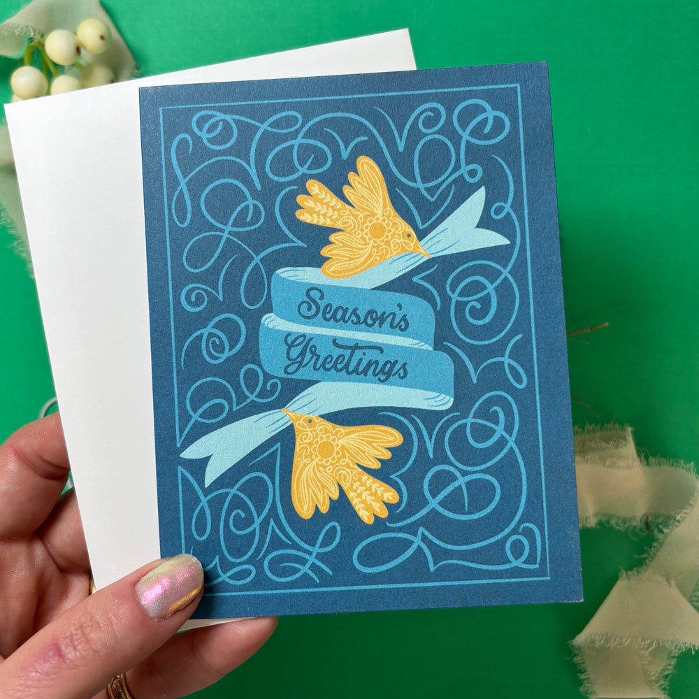 Hand holding a blue greeting card with yellow flowers and 'Season's Greetings' text on a green background.