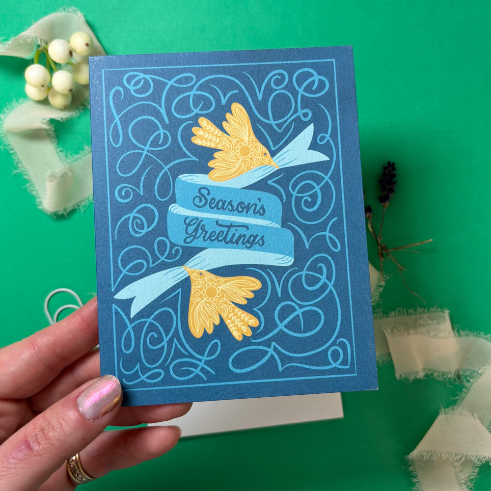 Blue greeting card with gold birds and 'Season's Greetings' text held by a hand on a green background.