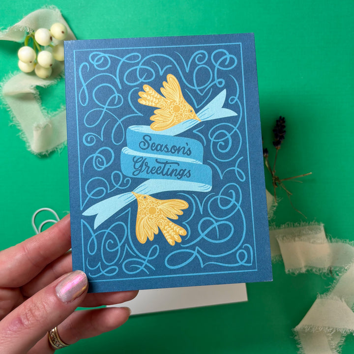 Blue greeting card with gold birds and 'Season's Greetings' text held by a hand on a green background.