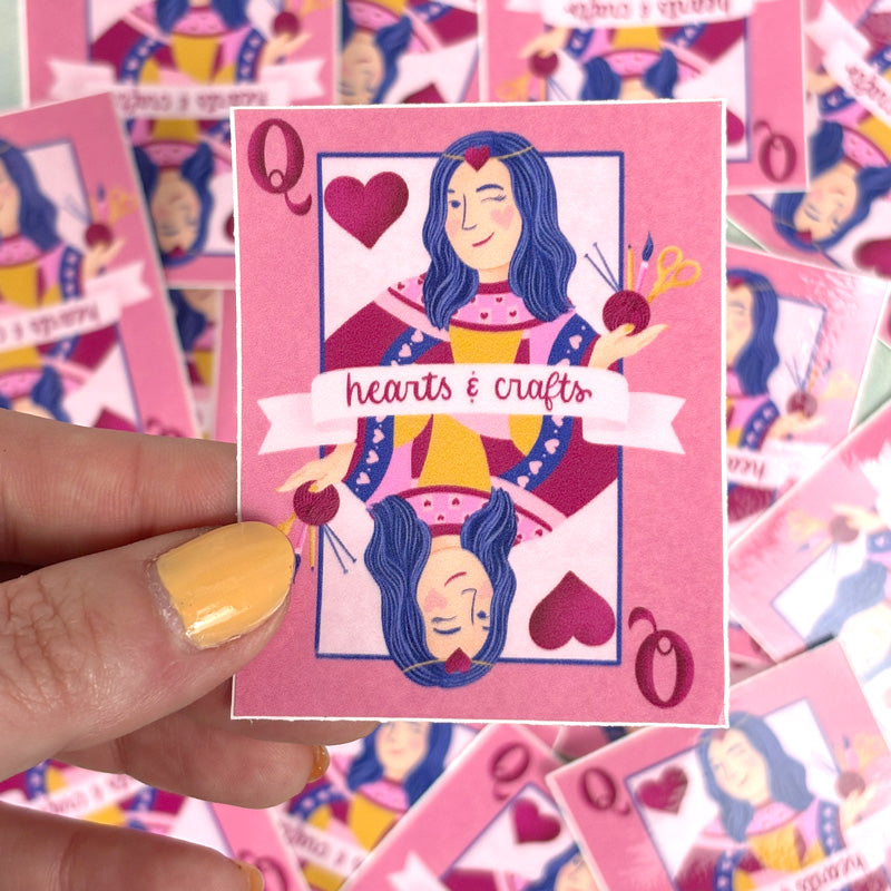 hand holding A pink sticker with a design of a queen playing card surrounded by hearts and crafting tools. The text 'HEARTS & CRAFT' is also visible.