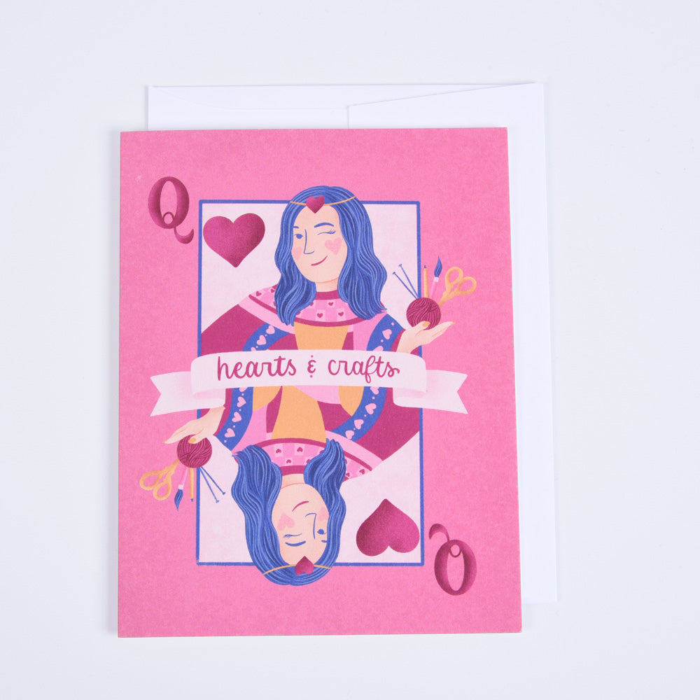 A folded greeting card with a pink background featuring an illustration of a woman with blue hair and hearts, and text that reads 'Hearts & Crafts'." The card is blank inside and has a bright white envelope.