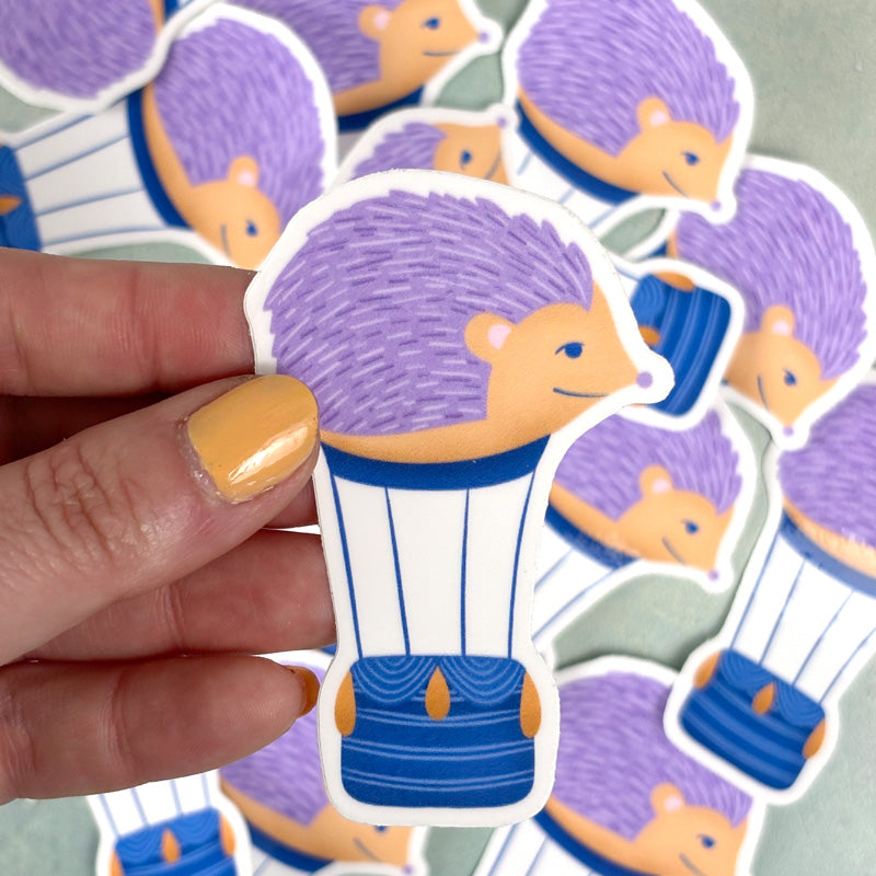 hand holding a sticker depicting a cartoon hedgehog riding a hot air balloon, with a blue and white color scheme and a purple hedgehog head.