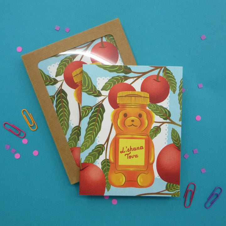 A set of greeting cards in a kraft box, styled on a bright blue background with paper clips and confetti. The card features an illustration of a bear-shaped honey pot with the text 'L'shana Tova' on a yellow label, surrounded by red apples and green leaves.
