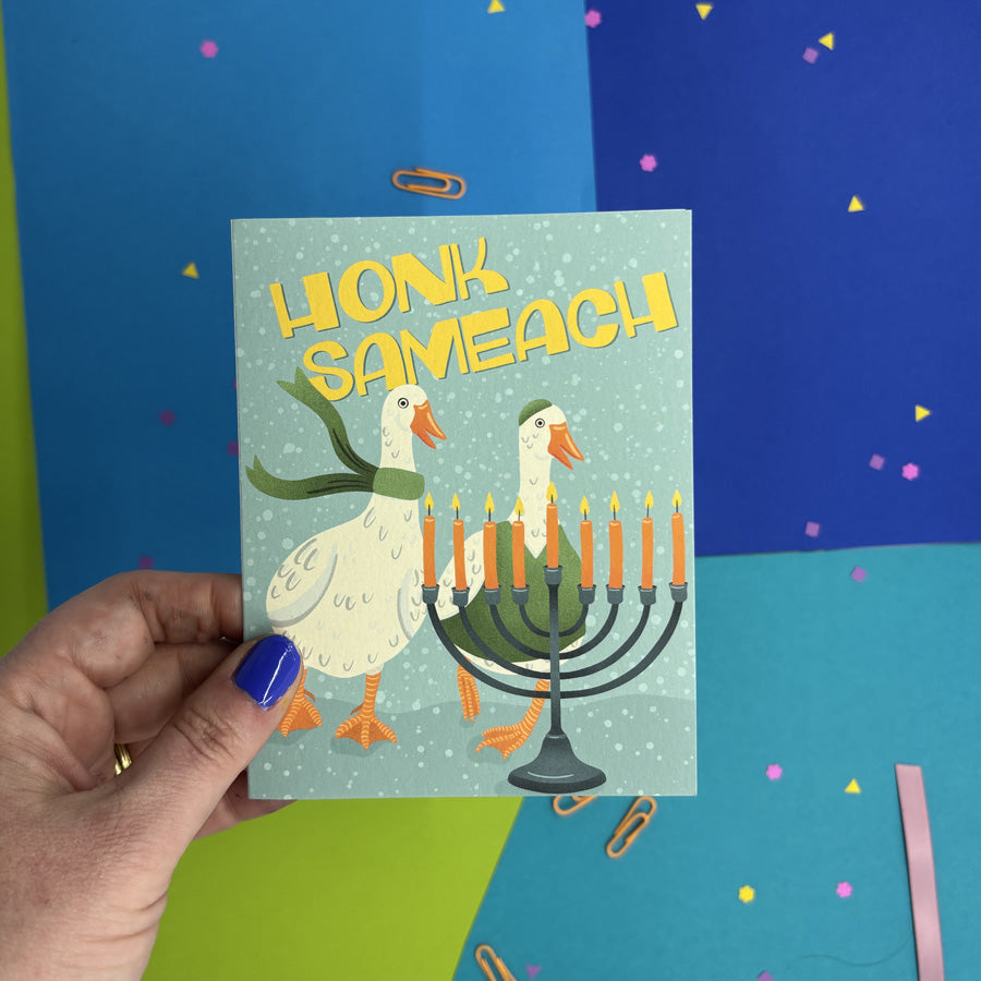 hand holding a hanukkah card featuring illustration of two geese wearing winter clothes standing behind a hanukkah menorah with text "honk sameach"