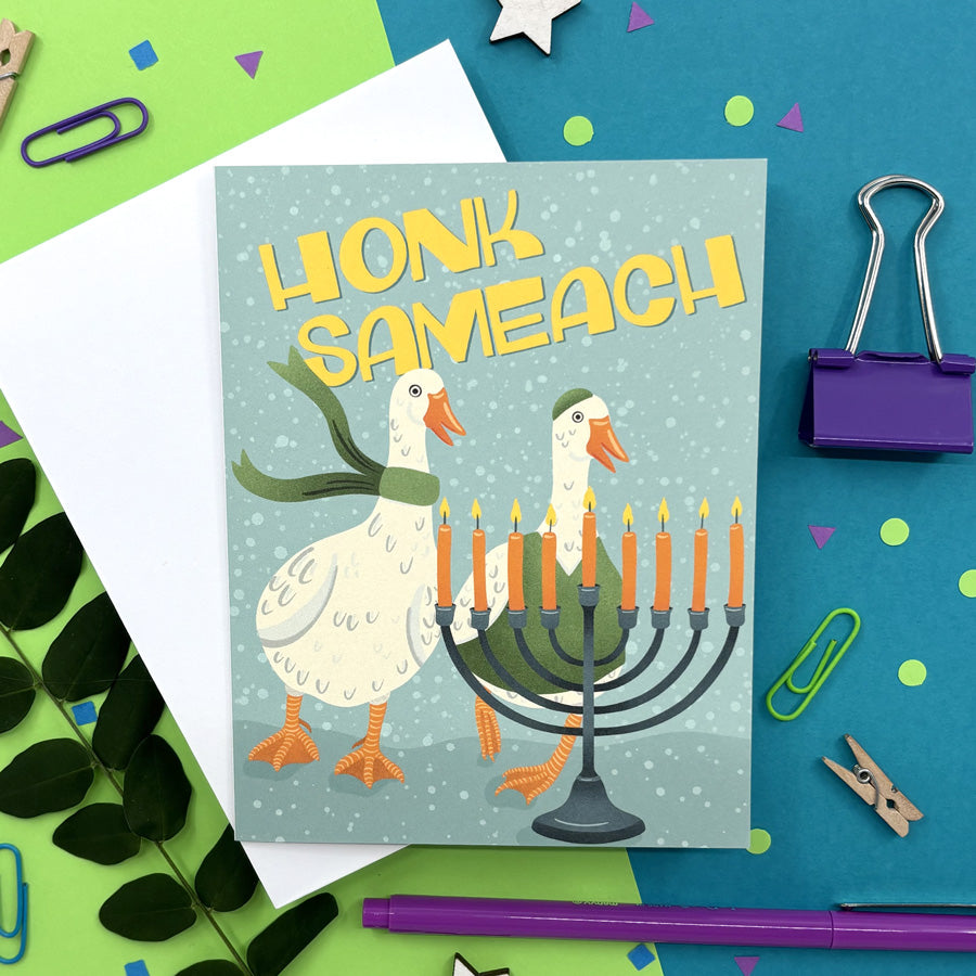 hanukkah card featuring illustration of two geese wearing winter clothes standing behind a hanukkah menorah with text "honk sameach" on a colorful background with stationery items