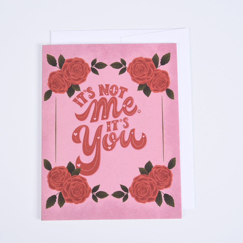 A pink greeting card with the text 'It's Not Me It's You' surrounded by red roses and green leaves, designed for an anti-Valentine's Day message.