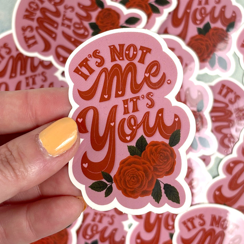 hand holding A pink sticker with the text 'It's Not Me' and an illustration of two red roses.