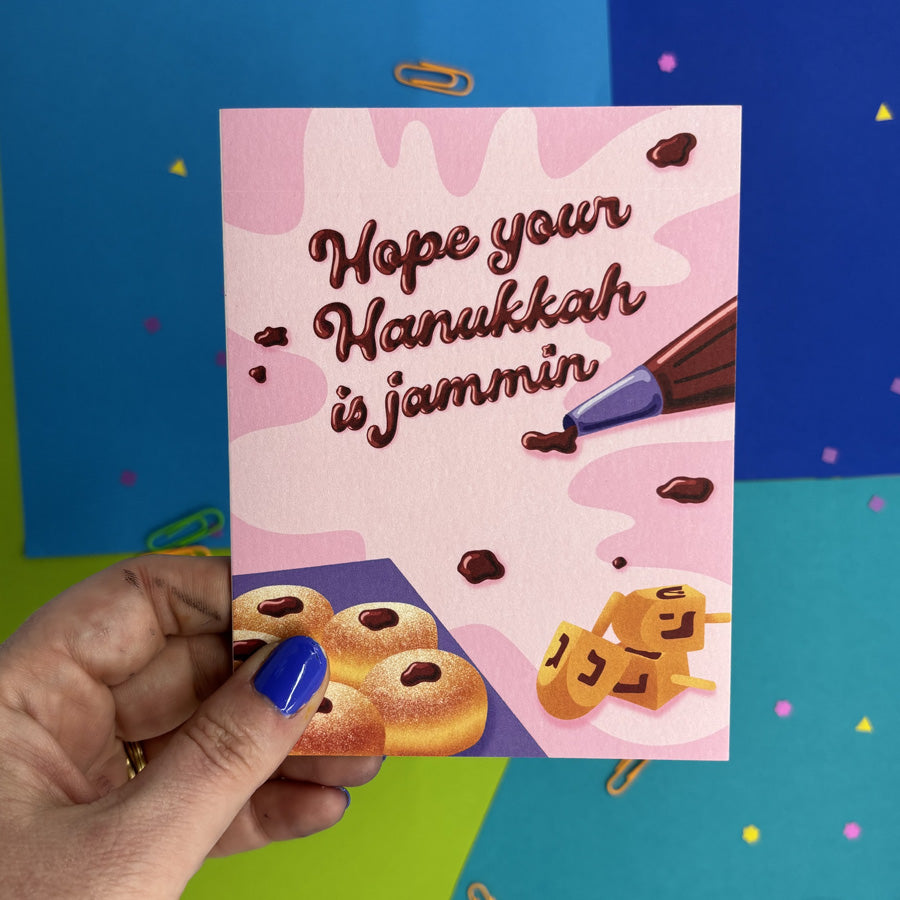 hand holding a hanukkah card featuring illustrated jelly donuts and piping bag with jam spelling "hope your hanukkah is jammin"