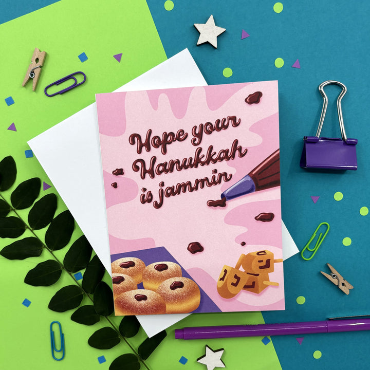 hanukkah card featuring illustrated jelly donuts and piping bag with jam spelling "hope your hanukkah is jammin" on a bright background with stationery items