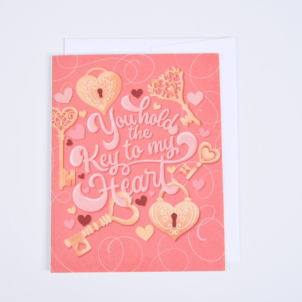 A pink folded greeting card with 'Key to My Heart' text and decorative elements such as hearts and a key motif, paired with a bright white envelope.