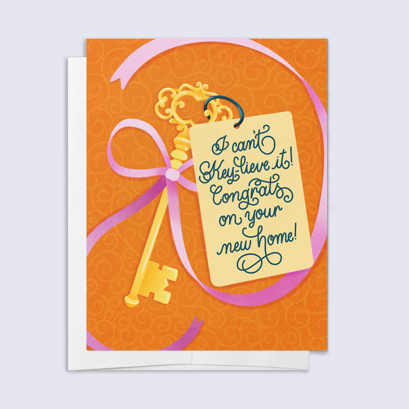 Greeting card with a gold key and pink ribbon on an orange background, featuring text 'I can't key lieve it! Congrats on your new home!'.
