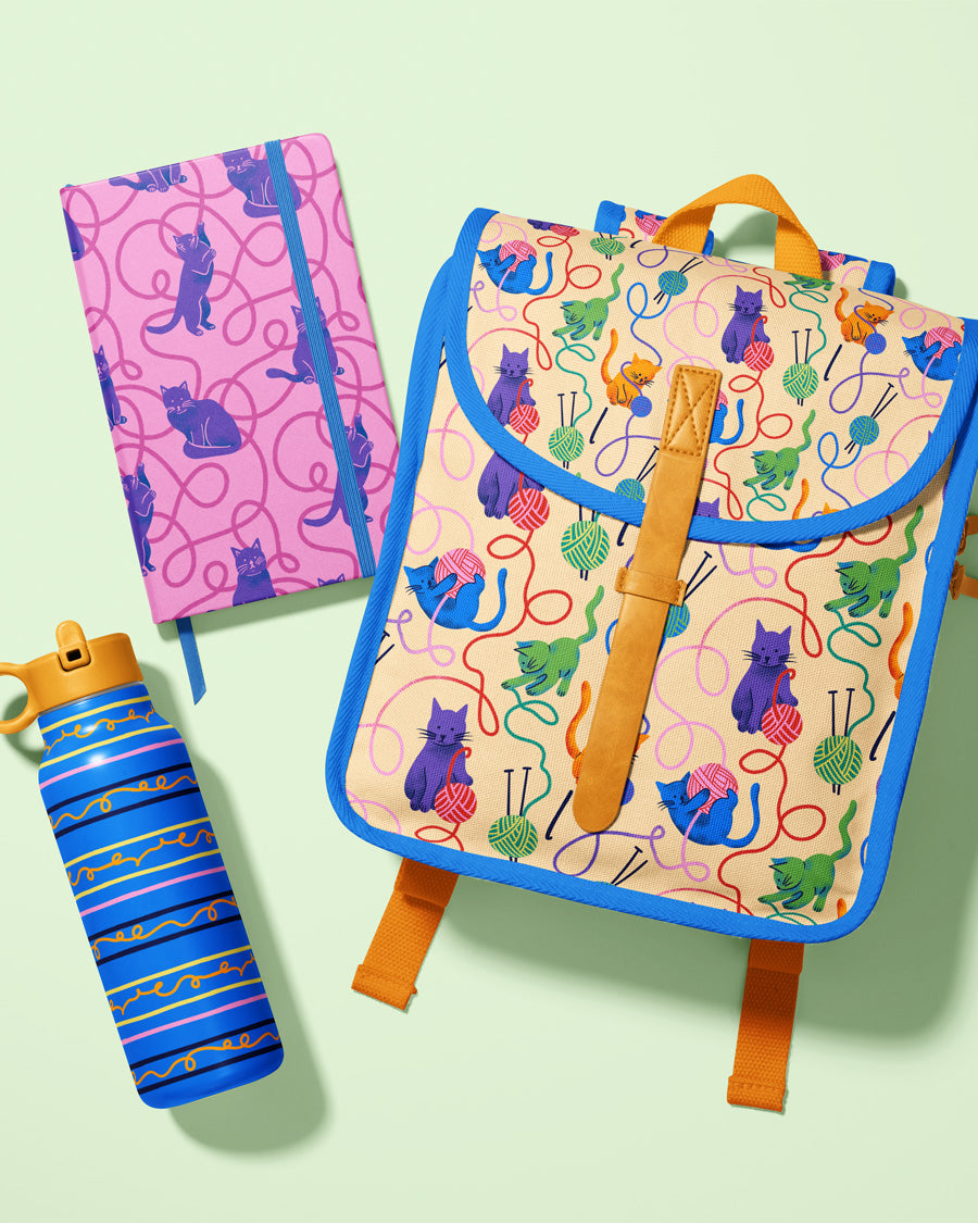 Colorful backpack with cat pattern, pink notebook, and blue striped water bottle on a light green background