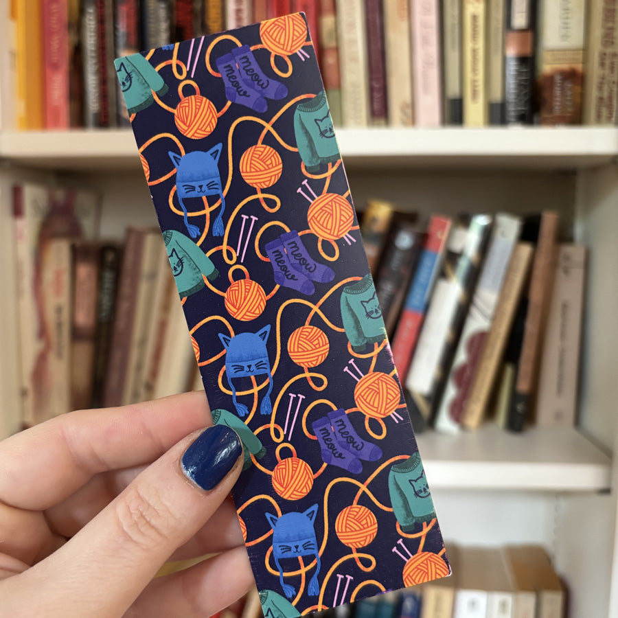 Hand holding a colorful bookmark with yarn and cat pattern in front of a bookshelf.