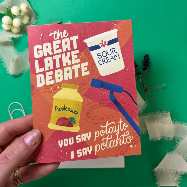 Card titled 'The Great Latke Debate' held by a hand on a green background with decorative elements.
