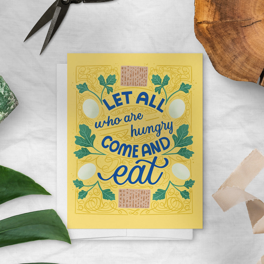 Yellow card with blue text 'Let all who are hungry come and eat' on a white surface with green leaves and scissors.