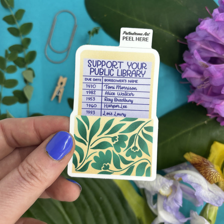 sticker with 'Support Your Public Library' text held by a hand against a floral and blue background
