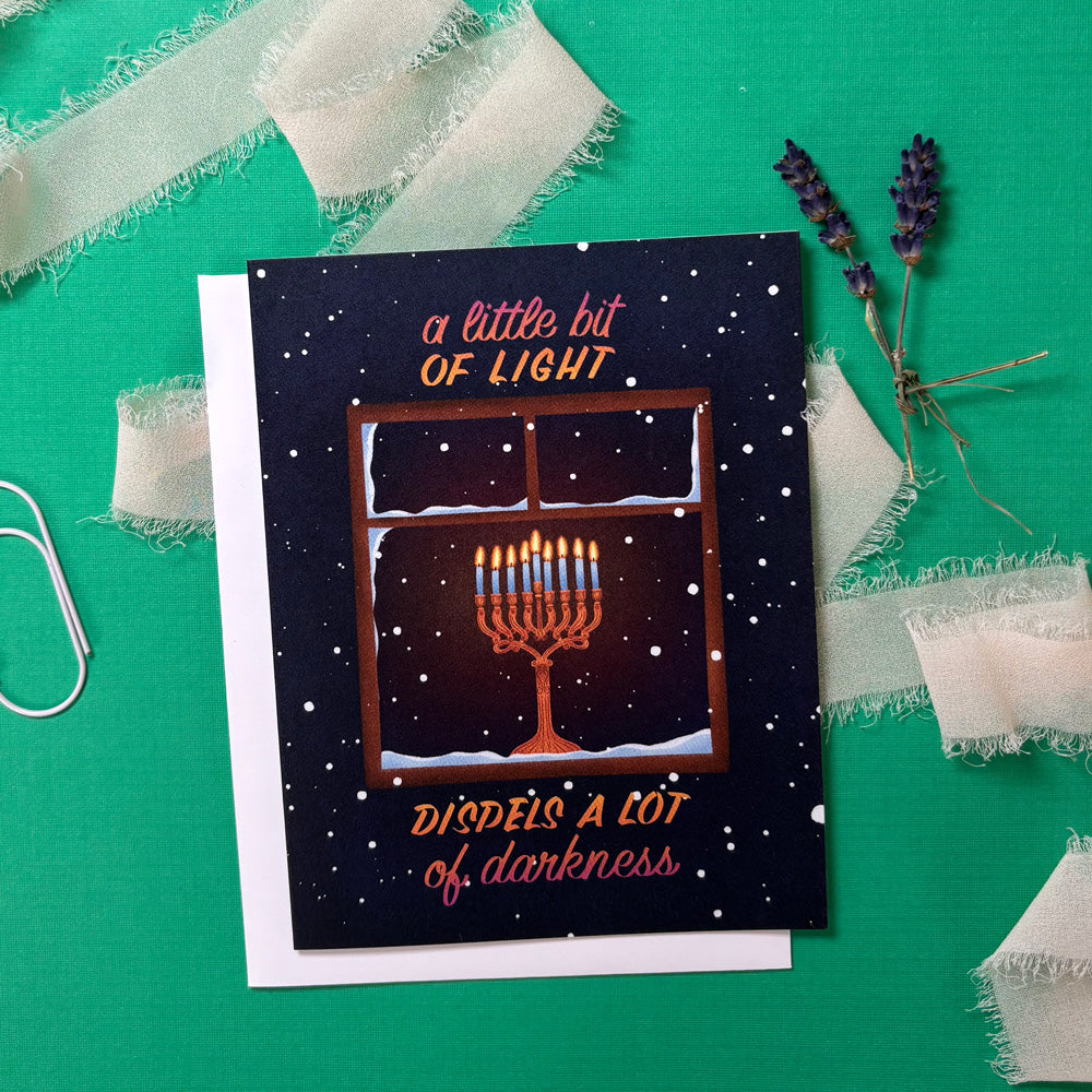 Greeting card with Menorah illustration and text "a little bit of light dispels a lot of darkness" on a green background