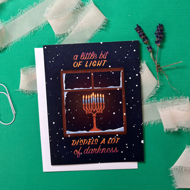 Greeting card with Menorah illustration and text "a little bit of light dispels a lot of darkness" on a green background