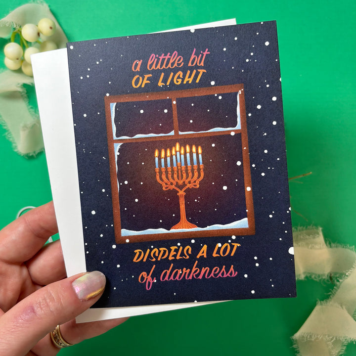 Card with Menorah illustration in a windowsill and text held by a hand on a green background