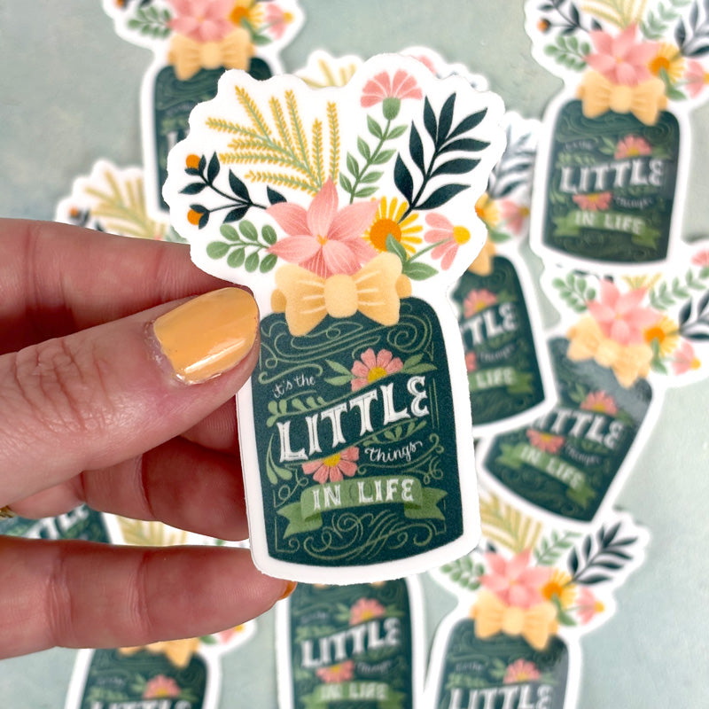 hand holding a sticker of a flower vase with lettering "it's the little things in life"