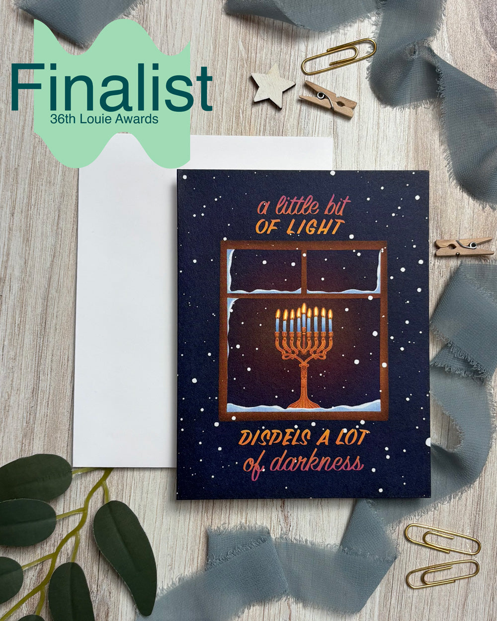 greeting card of a menorah in a window with text "a little bit of light dispels a lot of darkness" and a banner "finalist 36th Louie Awards"