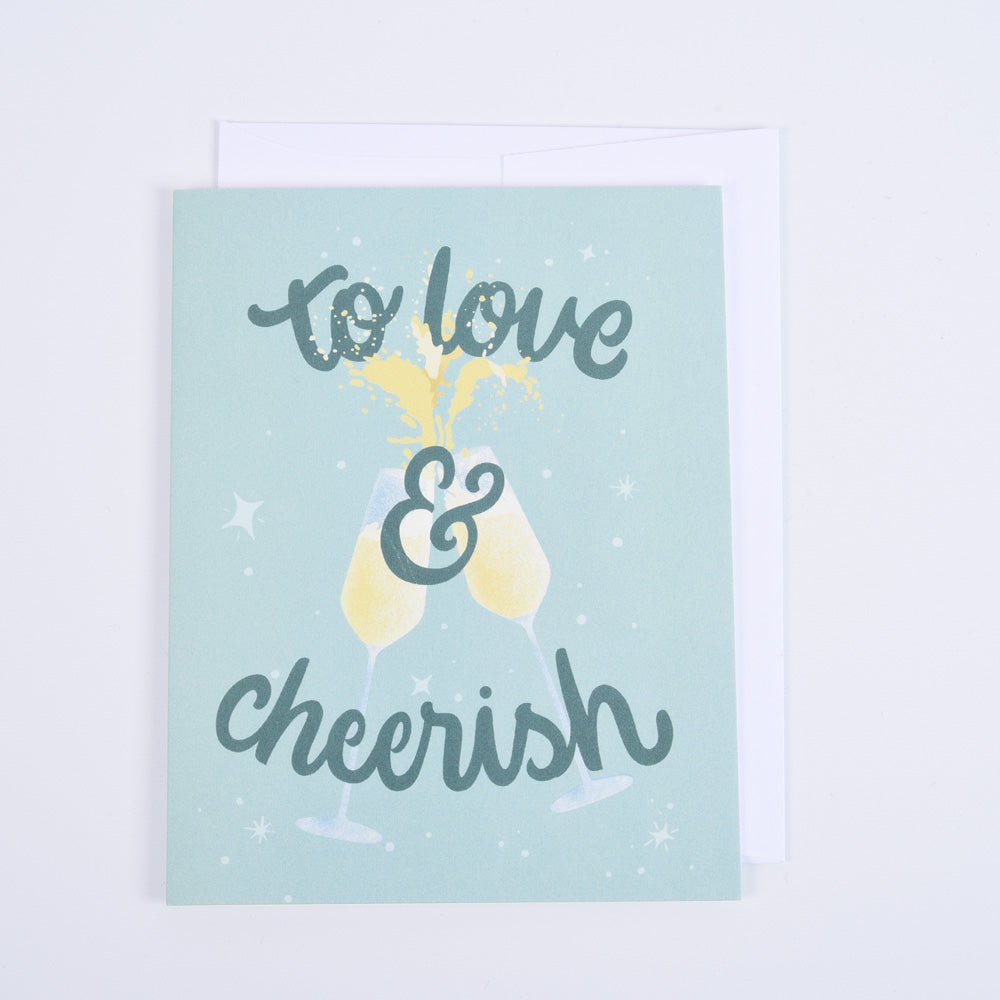 A folded greeting card with the text 'To Love & Cheerish' on a green and white background, featuring a champagne glass, and paired with a bright white envelope.