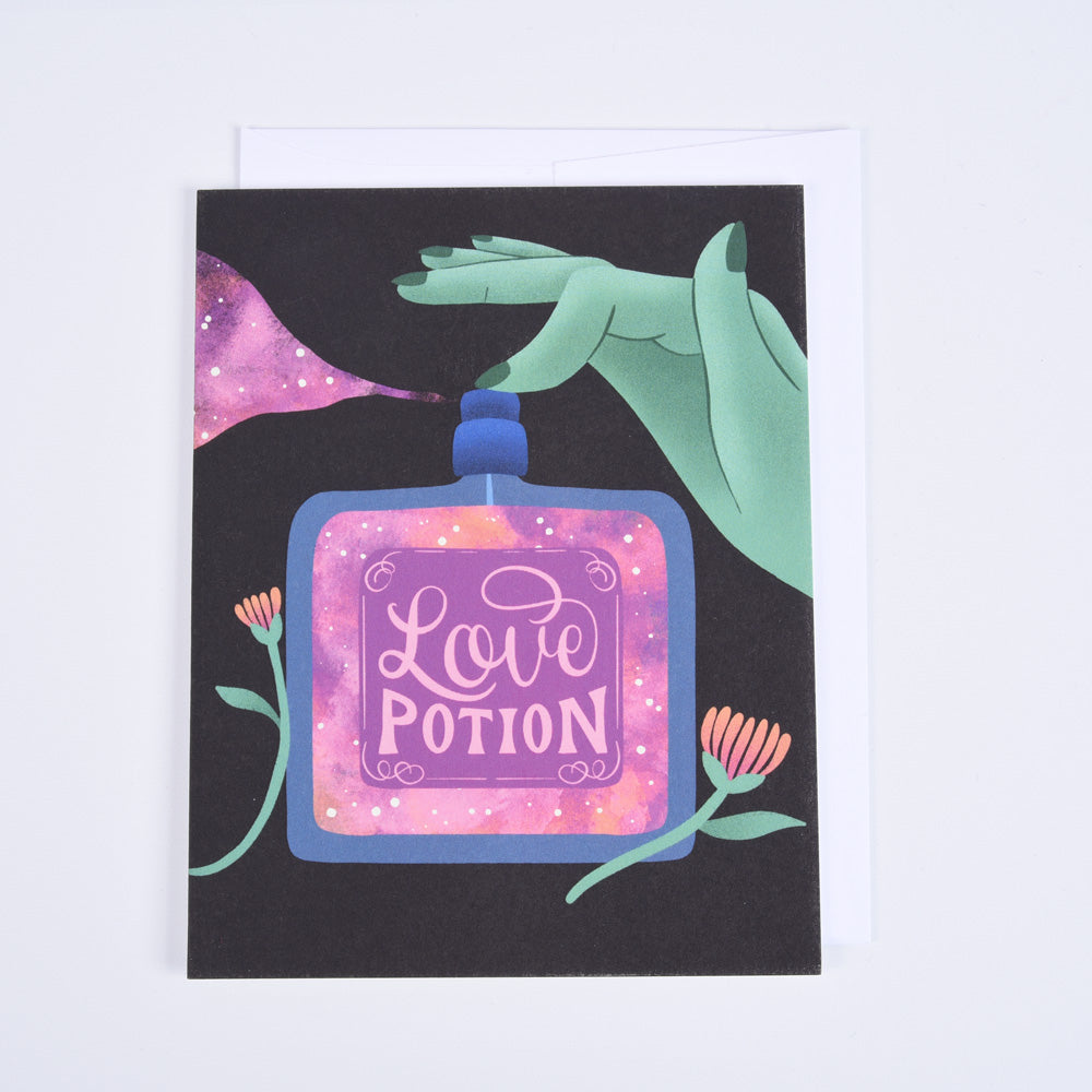 A folded greeting card with a colorful illustration of a love potion bottle, stars, and a green hand, with the phrase 'love potion' written on the front.