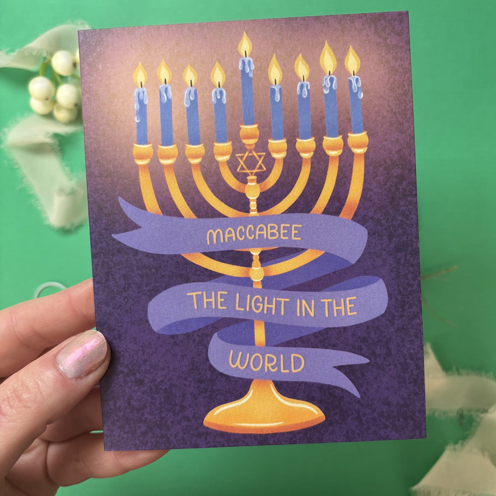 Card with Menorah design and text 'Maccabee, The Light in the World' held by a hand against a green background.