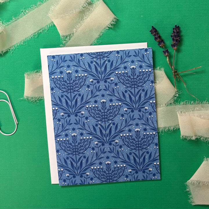 Blue floral menorah patterned card on a green background with fabric pieces and lavender.