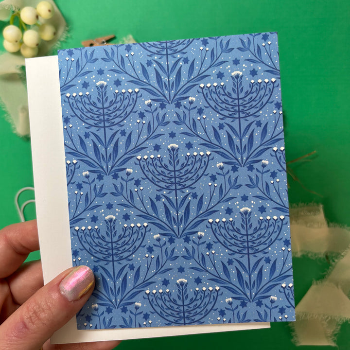 Blue patterned menorah card held by a hand against a green background