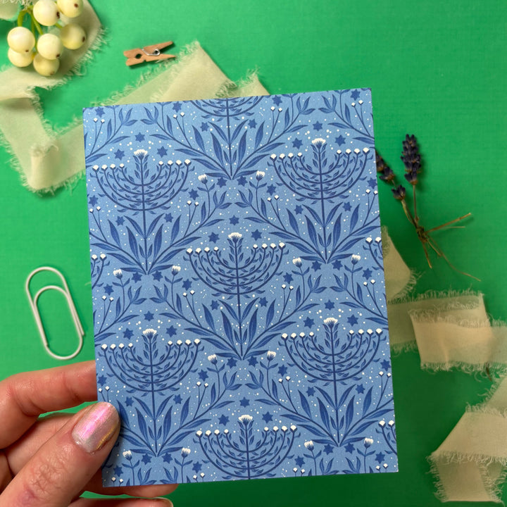 Blue patterned menorah card held by a hand on a green background with decorative items.