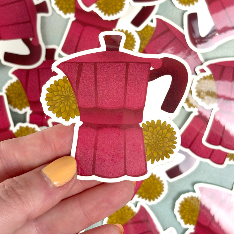 hand holding a sticker depicting a red Moka pot with yellow flowers 