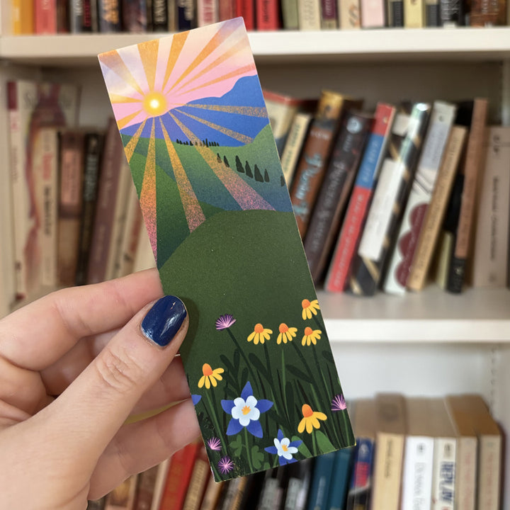 Hand holding a colorful bookmark with a nature design in front of a bookshelf.