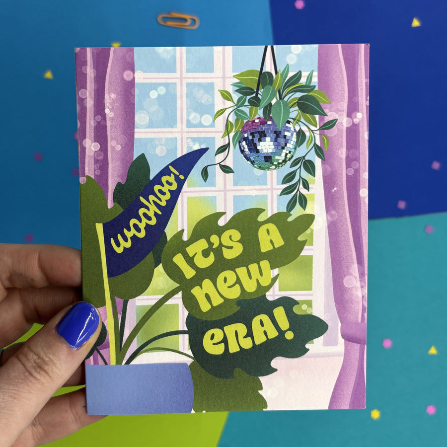 hand holding greeting card with an illustration of a disco ball hanging in front of an open window with a monstera plant and text "woohoo! it's a new era!"