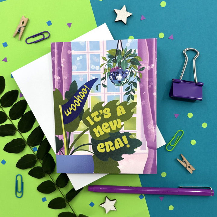 greeting card with an illustration of a disco ball hanging in front of an open window with a monstera plant and text "woohoo! it's a new era!" on a bright background with stationery items