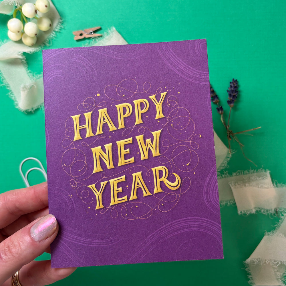 Purple 'Happy New Year' card held by a hand on a green background with decorative elements.