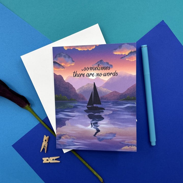 greeting card with illustration of a sun setting behind lake and mountains with a sailboat and script "sometimes there are no words" on a colorful background
