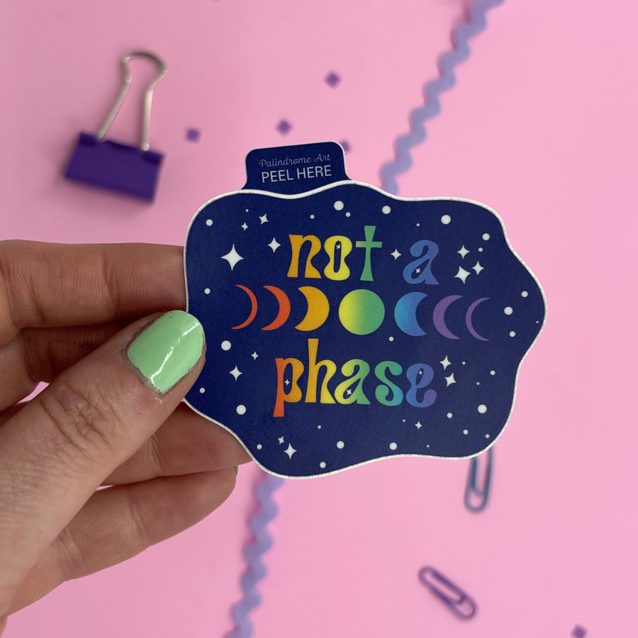 A hand with mint green nail polish holds a die-cut sticker that reads 'not a phase' in rainbow lettering on a dark blue starry background. The sticker has a small tab at the top reading 'Palindrome Art PEEL HERE'.