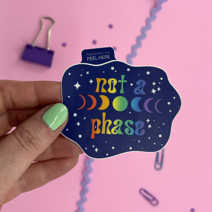 A hand with mint green nail polish holds a die-cut sticker that reads 'not a phase' in rainbow lettering on a dark blue starry background. The sticker has a small tab at the top reading 'Palindrome Art PEEL HERE'.