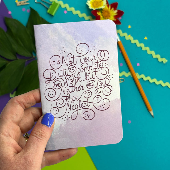 A hand with blue nail polish holds a small notebook with a lavender watercolor cover and cursive text that reads, 'It is not your duty to work but neither are you free to neglect it.' The background is a bright teal surface with decorative paper clips, a pencil, green zigzag paper, and flowers.