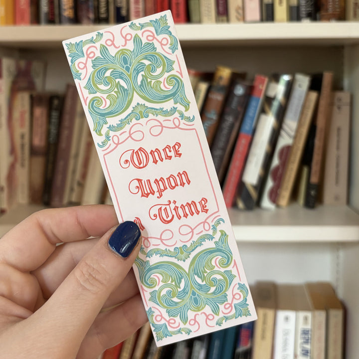 Hand holding a decorative bookmark with 'Once Upon a Time' text in front of a bookshelf.