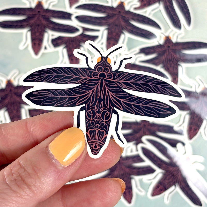 hand holding A sticker of a purple open-winged beetle with a floral and garden-themed design.