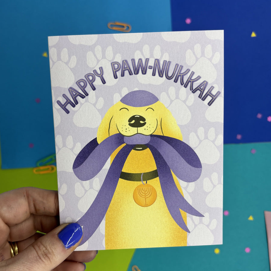 Hand holding a hanukkah card with a cute illustrated dog wearing a kippah and menorah collar holding a big gift bow in its mouth with text "happy paw-nukkah"