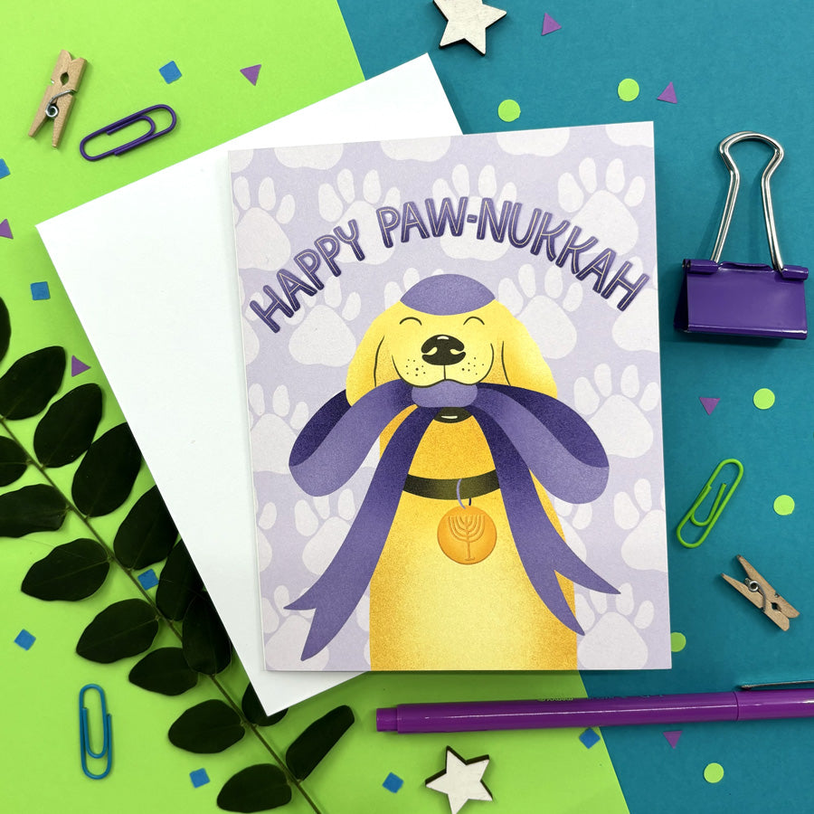 hanukkah card with a cute illustrated dog wearing a kippah and menorah collar holding a big gift bow in its mouth with text "happy paw-nukkah" on a colorful background ith stationery items