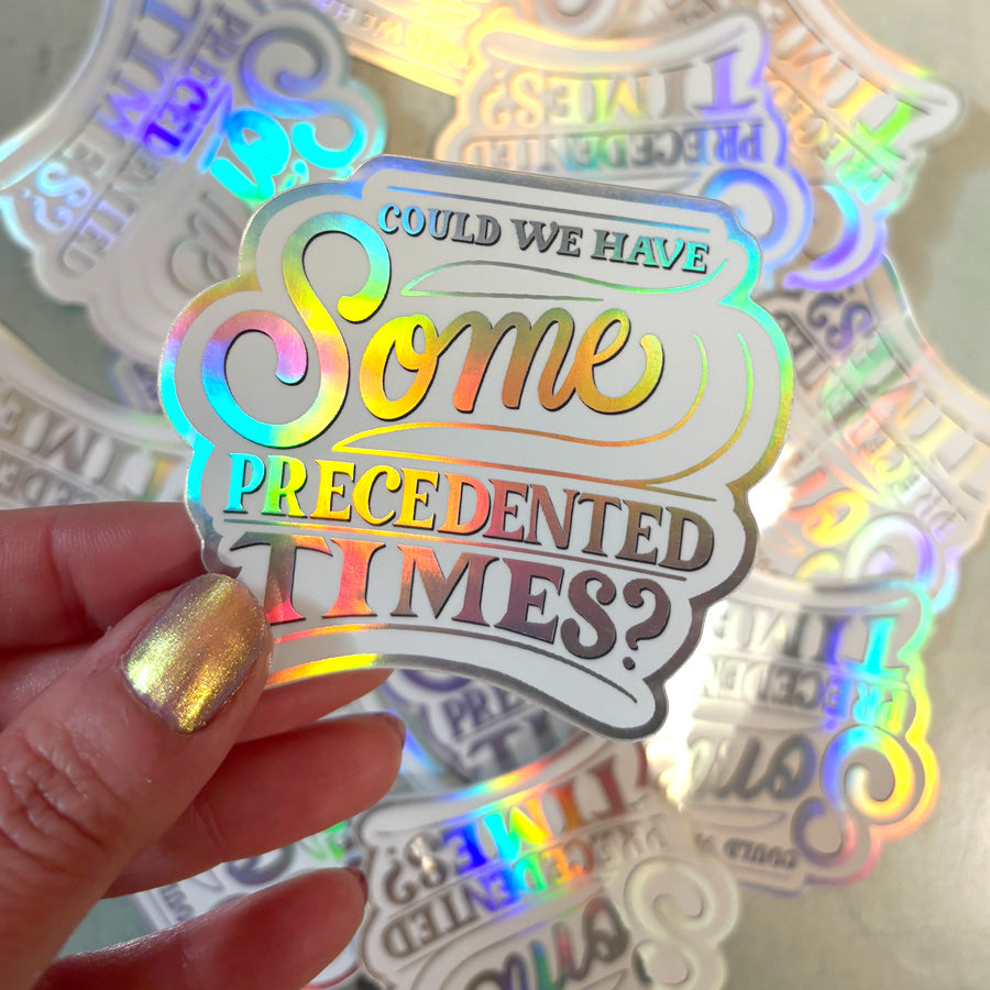 Holographic sticker with colorful text that says "could we have some precedented times?"  held by a hand