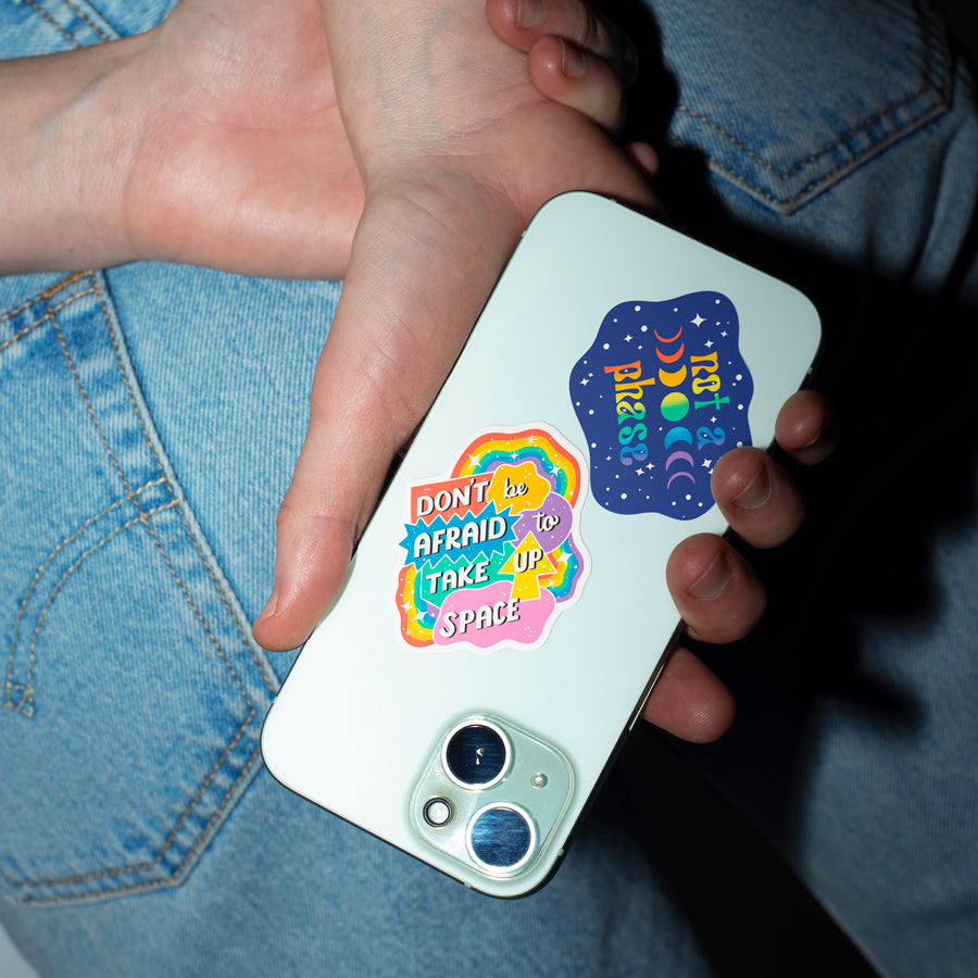 A person holding a white smartphone case with two colorful retro-style stickers. One sticker reads 'DON'T BE AFRAID TO TAKE UP SPACE' in rainbow colors, and the other reads 'IT'S OK TO MISS' in blue with yellow and orange moon and star graphics.