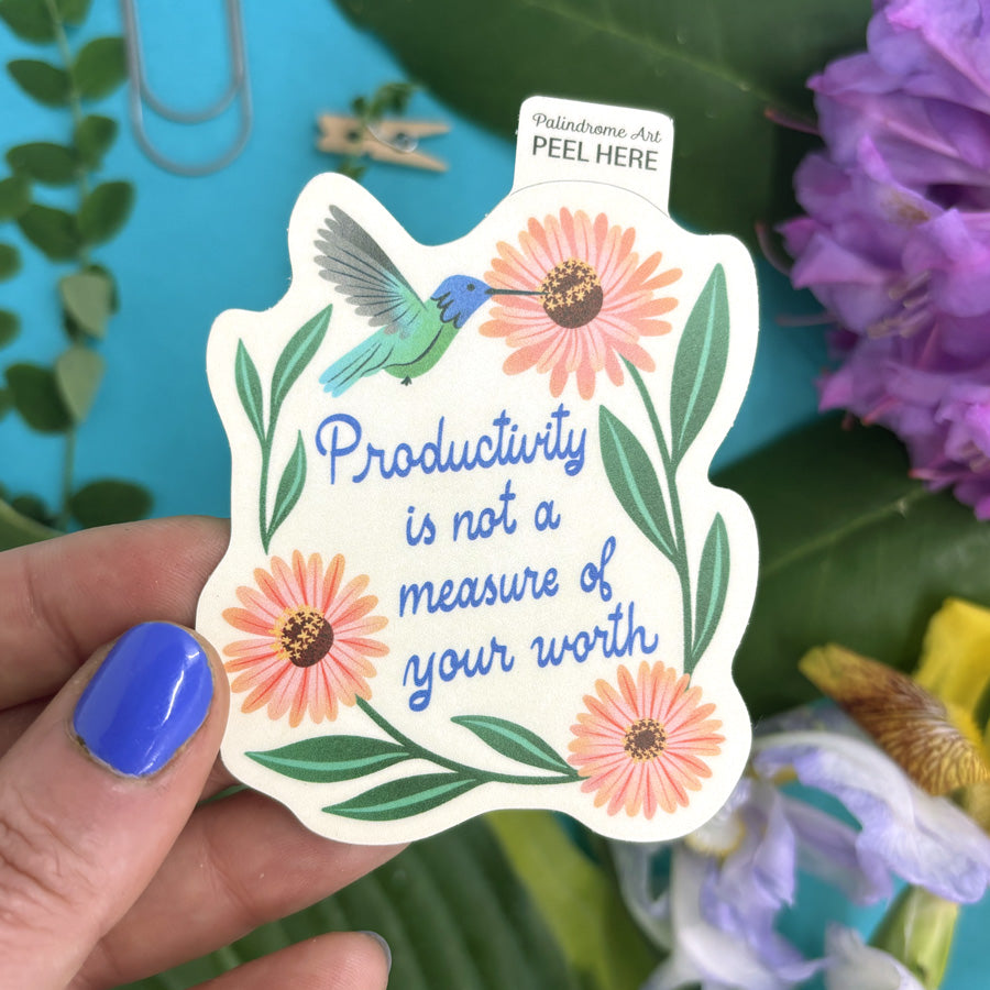 Hand holding a sticker with a hummingbird, flowers, and text "productivity is not a measure of your worth"