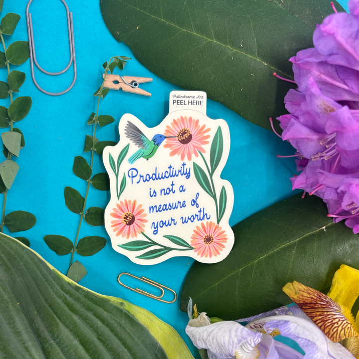 Decorative sticker with a hummingbird, flowers, and text "productivity is not a measure of your worth" on a bright background with flowers and stationery items