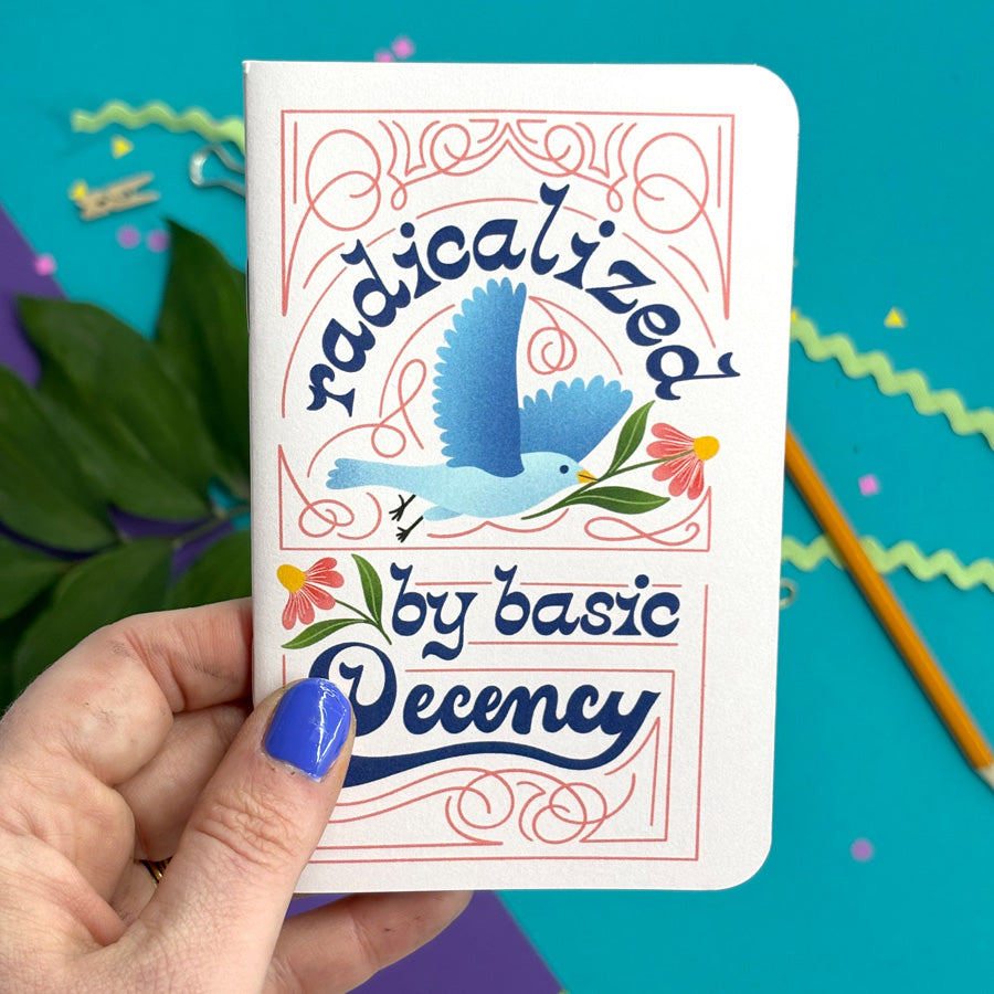A hand with blue nail polish holds A small pink notebook with rounded corners featuring a colorful retro-style illustration of a blue bird and the text 'radicalized by basic decency' in dark blue script. The background is a bright teal surface with decorative paper elements.