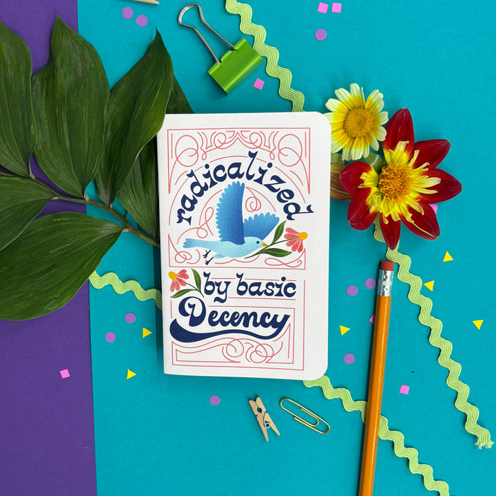 A small pink notebook with rounded corners featuring a colorful retro-style illustration of a blue bird and the text 'radicalized by basic decency' in dark blue script is arranged in a styled flat lay on a bright turquoise background with purple paper, green leaves, flowers, a pencil, binder clips, and paper clips.